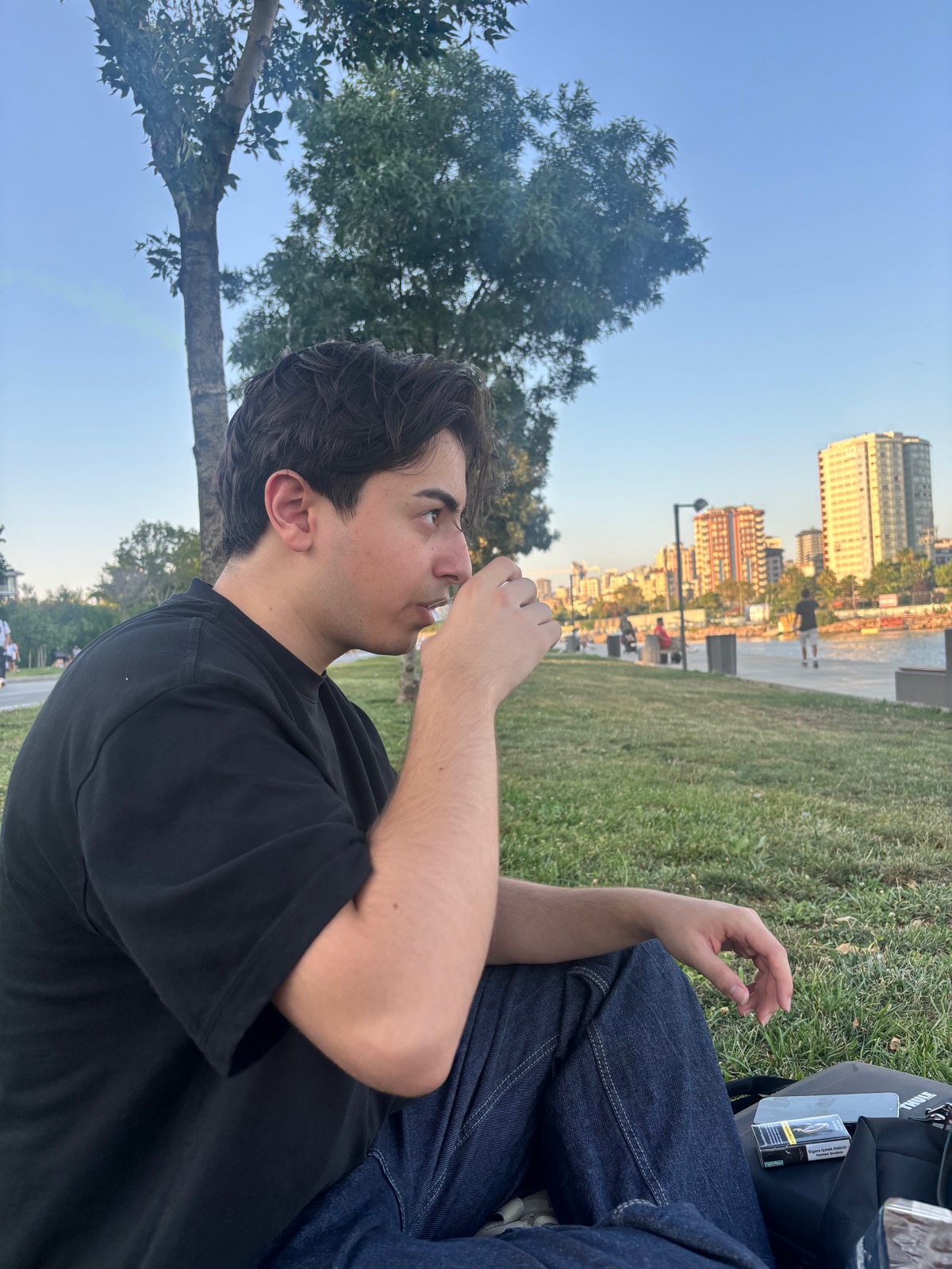 A young man in conversation sitting on the grass along a waterfront promenade in Istanbul, drinking from a small cup with city buildings across the water.