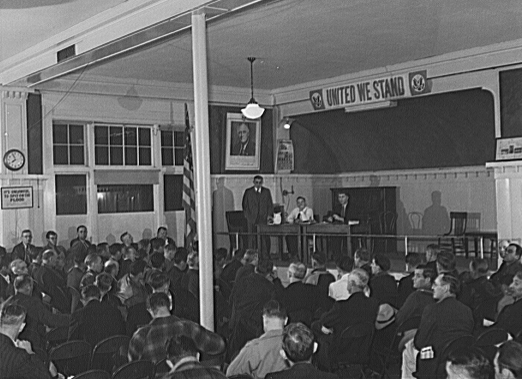 An audience of miners seated in an auditorium listening attentively to a speaker on a raised stage under a banner reading 'United We Stand' in Butte, Montana, 1942