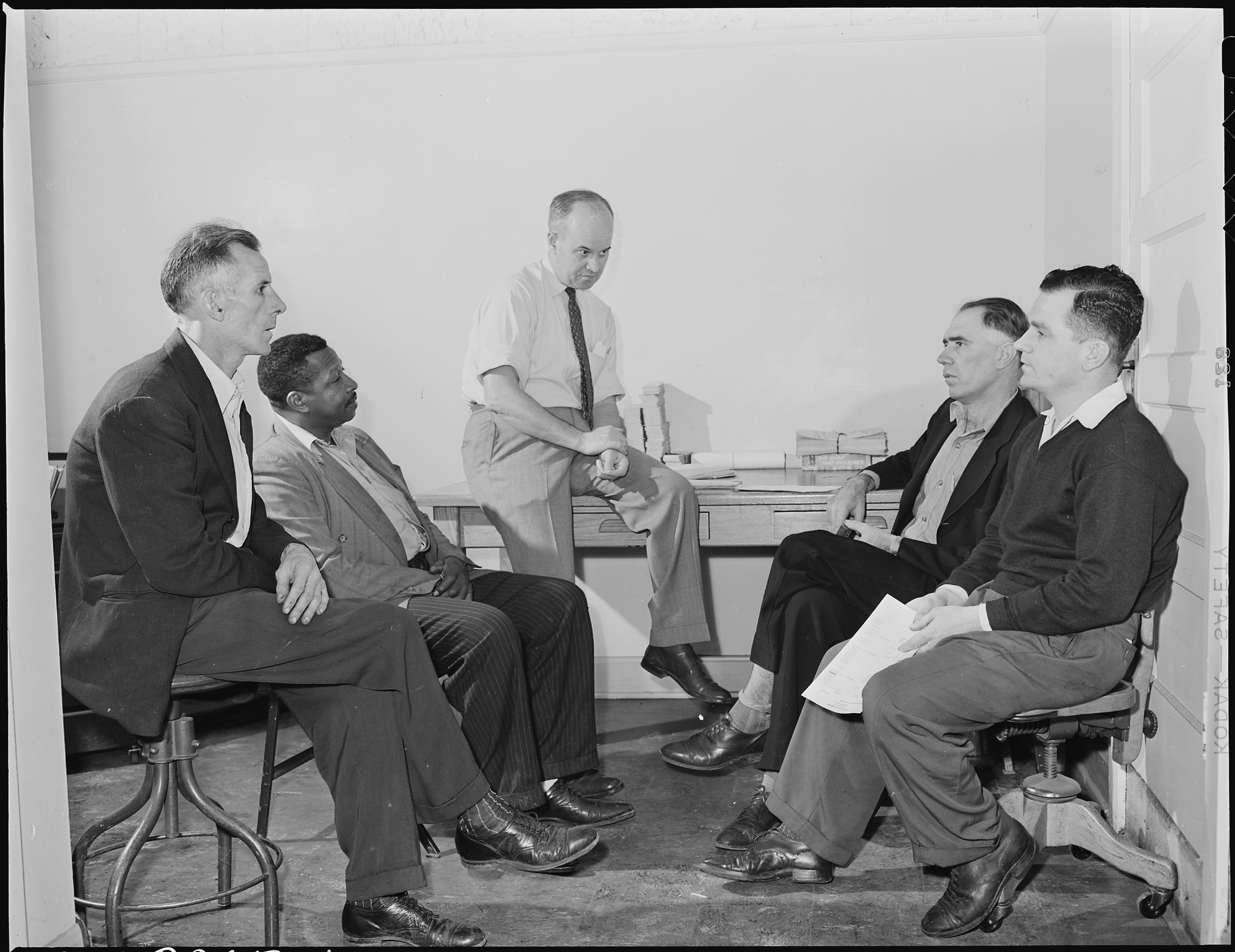 Five men in a 1946 association meeting seated closely around an office desk, listening intently during a discussion in Wheelwright, Kentucky.